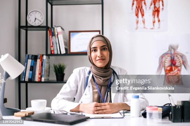 smiling asian muslim woman doctor is wearing a lab coat, hijab and a stethoscope sitting by the table in doctor's office. - arabic doctor stockfoto's en -beelden