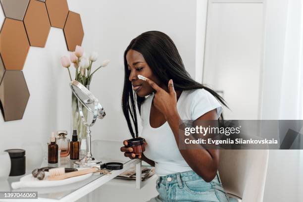 beautiful young black woman holding facial cream bottle and applying it at home. - dressing table stock pictures, royalty-free photos & images