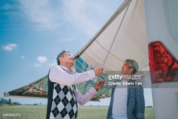 2 asian chinese senior man setting up awning canopy of campervan motor home during day time beside rice paddy field
