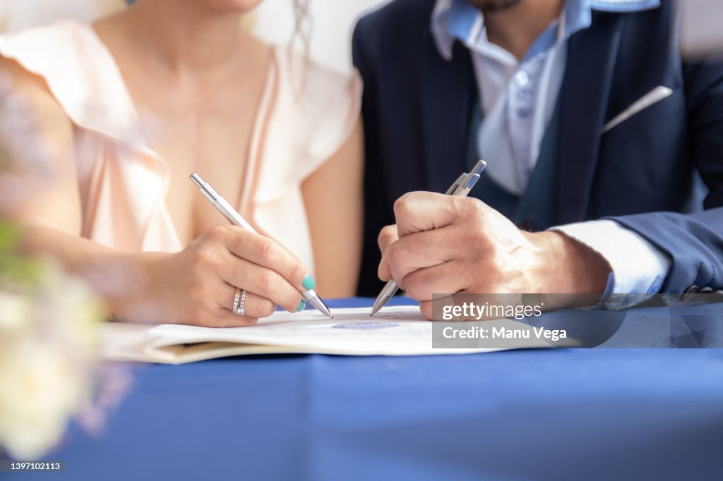 Close-up view of man and woman signing marriage certificate, she wears a vintage wedding dress and he wears a formal suit.