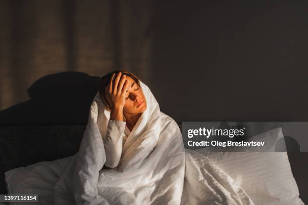 woman has a headache and sitting in bed covered with blanket. high temperature, virus or flu - awake stock pictures, royalty-free photos & images