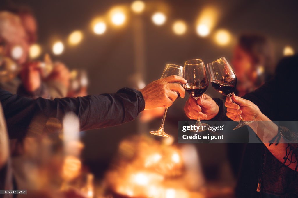 Celebratory red wine toast between senior adult friends at candle light social event party with string fairy lights