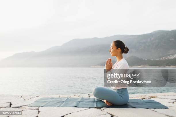 young woman praying and meditating outdoors by seaside. self care and mindfulness, menthal health. awakening in morning - yoga stock-fotos und bilder