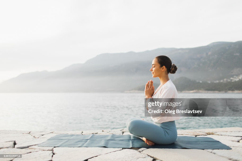 Young woman praying and meditating outdoors by seaside. Self care and mindfulness, menthal health. Awakening in morning