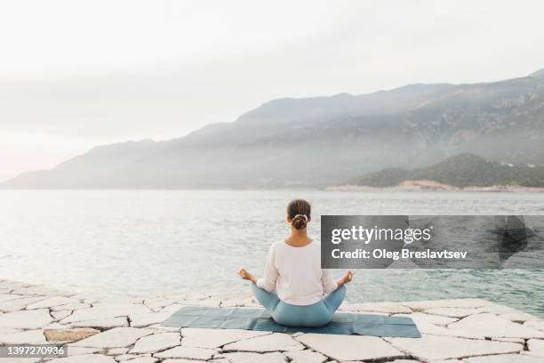 view from behind of woman in lotus position meditating outdoors with tranquil sea view. background with copy space. unrecognizable person - lotus position stock pictures, royalty-free photos & images