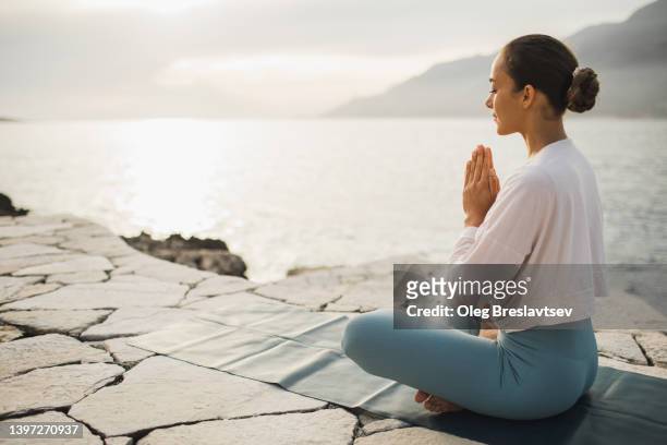 young woman praying and meditating outdoors by seaside. self care and mindfulness, menthal health. awakening in morning - lotus position stock pictures, royalty-free photos & images