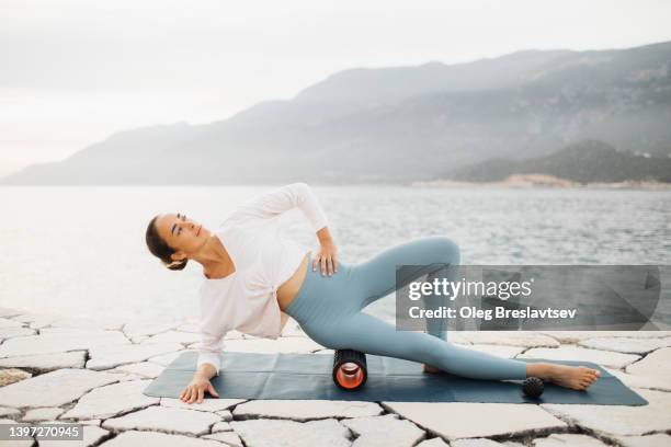 pretty young woman practicing self massage outdoors on yoga mat. massage roll on hips. cellulite prevention - hip stretch stock pictures, royalty-free photos & images