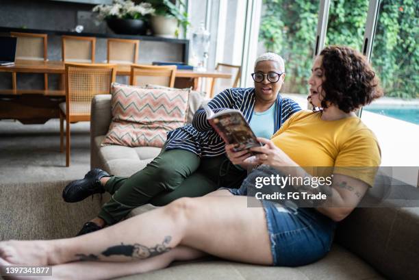 mid adult woman talking with a friend and reading a book at home - book people stock pictures, royalty-free photos & images