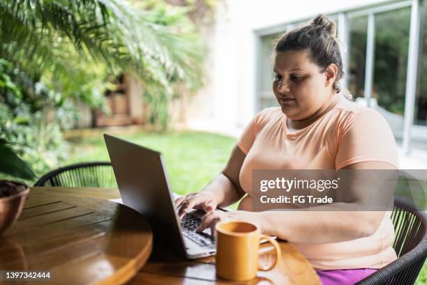 porträt einer mittelgroßen frau, die zu hause am laptop arbeitet - adipositas stock-fotos und bilder
