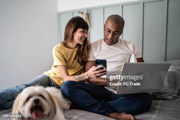 junge frau, die dem ehemann etwas auf dem handy mit dem hund im bett zu hause zeigt - couple looking laptop stock-fotos und bilder