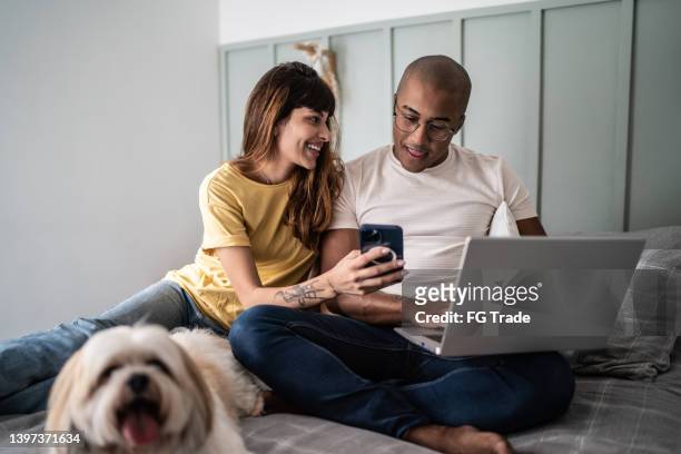 young woman showing something to husband on the mobile phone with the dog in the bed at home - telecommuting couple stock pictures, royalty-free photos & images