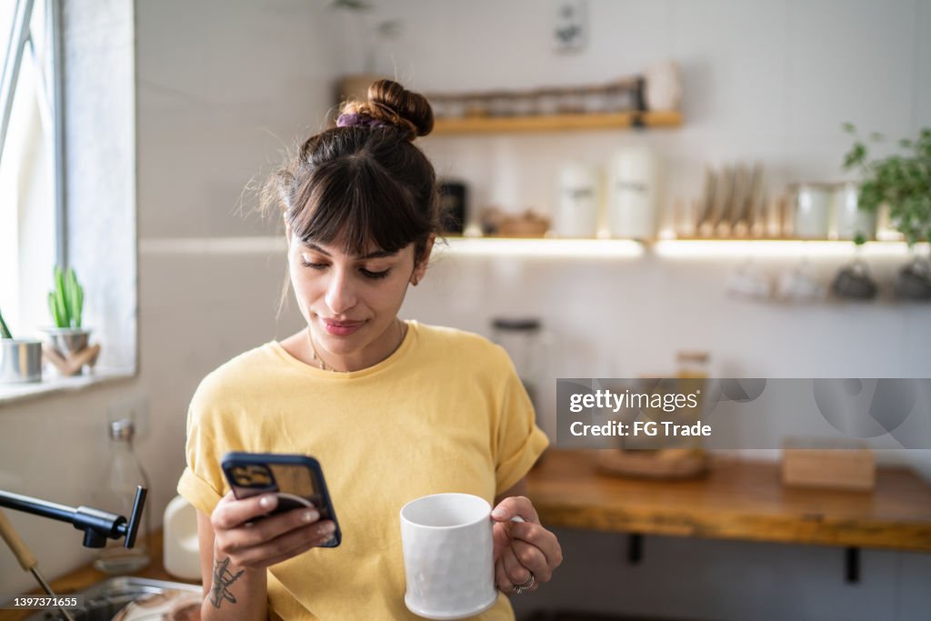 Junge Frau, die das Handy benutzt, während sie zu Hause Kaffee oder Tee trinkt