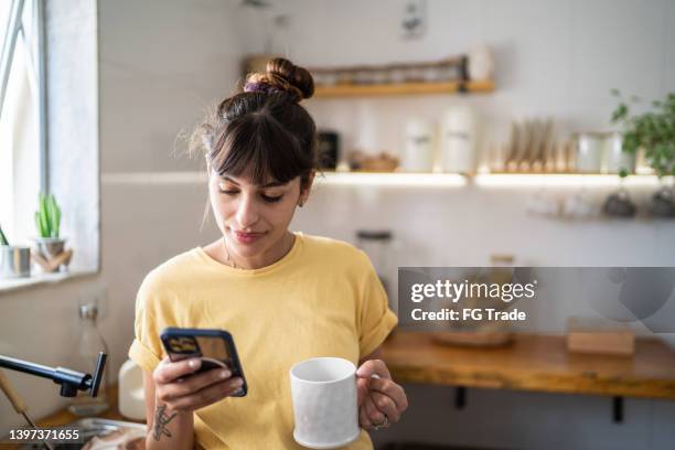 junge frau, die das handy benutzt, während sie zu hause kaffee oder tee trinkt - kitchen coffee home stock-fotos und bilder