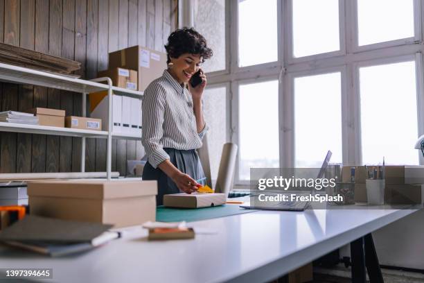 a happy businesswoman talking on her smartphone while preparing packages for shipping  in her store - stationary stock pictures, royalty-free photos & images