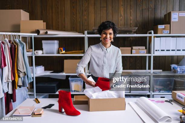 a smart businesswoman looking at camera while packing a pair of red boots for shipping - smart shoes stock pictures, royalty-free photos & images