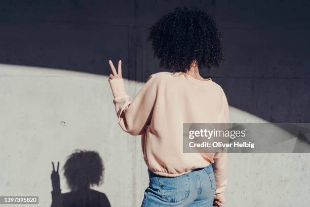 rear view of a young woman making a peace sign. on the wall you can see the shadow of the woman - sweatshirt stock-fotos und bilder