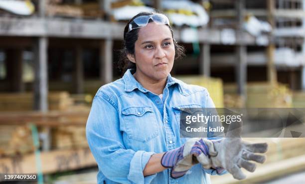 reife hispanische frau auf holzplatz zieht arbeitshandschuhe an - toughness stock-fotos und bilder