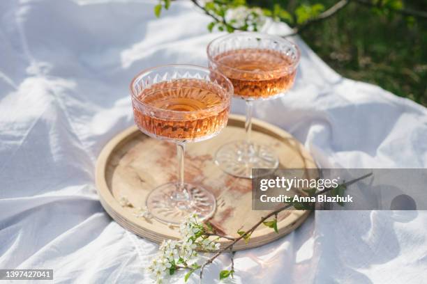 two glasses of rose wine outdoors on wooden tray and white textile, picnic in spring. sunny day, green grass and blossoming cherry trees. - green which rose stockfoto's en -beelden