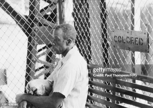 Man, wearing a white short-sleeve short, against a background of chainlink fencing on which is a sign reading 'Colored', United States, circa 1955.