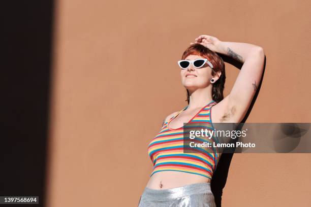 portrait of a non-binary young with sunglasses smiling and resting against a brown wall - armpit imagens e fotografias de stock