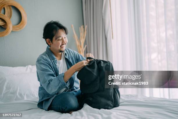 hipster asiatischer mann, der seinen rucksack (rucksack) in seinem schlafzimmer packt, um sich auf den weggang vorzubereiten. - man in suite holding tablet stock-fotos und bilder