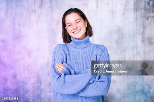 beautiful young woman with short hair and gray eyes laughing against gray wall background with crossed arms. she is wearing a violet sweater. concept of natural beauty - cuello alto fotografías e imágenes de stock