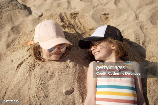 six year old girls laughing together on the beach, one girl has her body buried in the sand and the other girl is laying next to her friend - family summer holiday stock-fotos und bilder