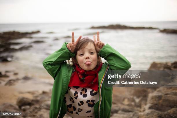 a 6 year old girl dressed with a green raincoat , posing and making funny face on rocks at the seaside - landskap stockfoto's en -beelden