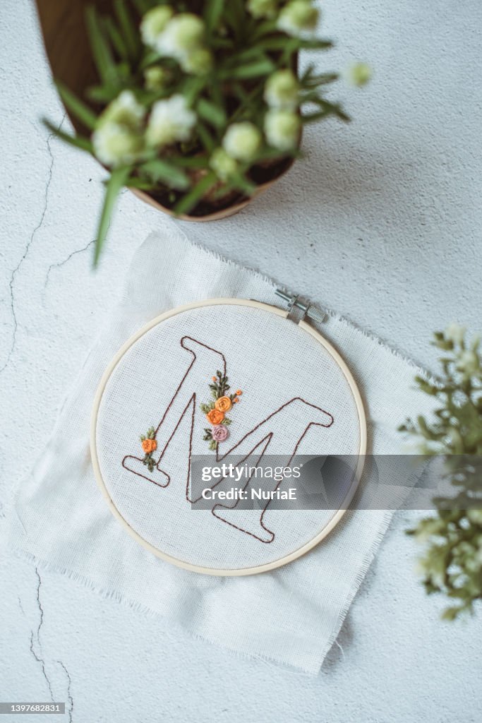 Overhead view of an embroidery hoop with the letter M on a table next to a bunch of flowers