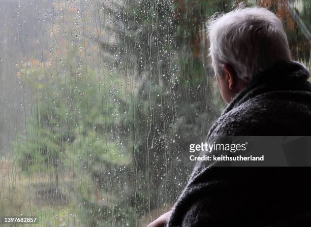 man wrapped in a blanket looking out of a window on a rainy day - window rain stock-fotos und bilder