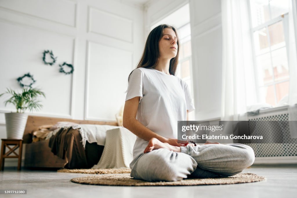 A young beautiful woman is sitting in a bright room on a jute rug and meditating with her eyes closed