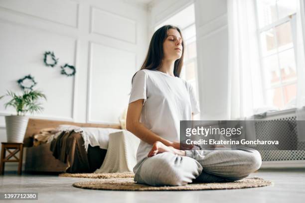 a young beautiful woman is sitting in a bright room on a jute rug and meditating with her eyes closed - salle yoga photos et images de collection