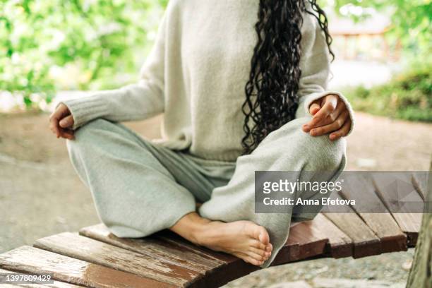 barefoot authentic woman with long curly hair and in cozy knitted clothing is sitting on a bench in lotus position. she is meditating at a public park. concept of relaxation exercises - grey trousers stock-fotos und bilder
