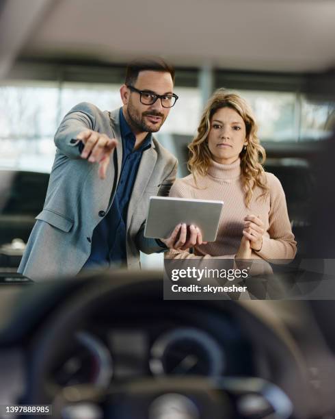 young car salesman showing a car to his customer in a showroom. - car inside showroom stock pictures, royalty-free photos & images