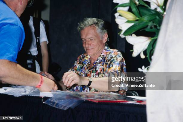 American magazine publisher Hugh Hefner , wearing a shirt featuring a collage of Playboy magazine covers, signing Playboy memorabilia at the Playboy...