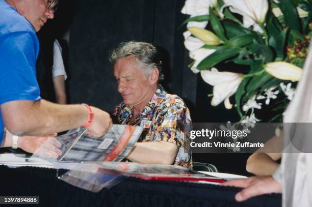 American magazine publisher Hugh Hefner , wearing a shirt featuring a collage of Playboy magazine covers, signing Playboy memorabilia at the Playboy...