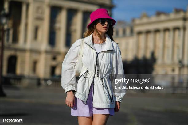 Maria Rosaria Rizzo wears a pink fuchsia bob hat from Kangol, gray sunglasses, silver earrings, a white t-shirt, a white zipper rain coat, pale...