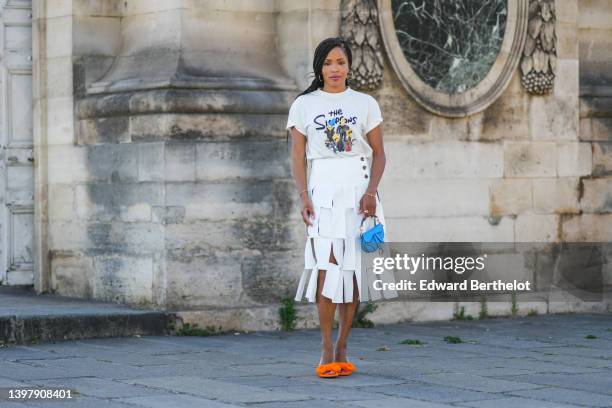 Ellie Delphine wears brown sunglasses, gold earrings, a white with black and blue and yellow t-shirt with The Simpsons print pattern, gold bracelets,...