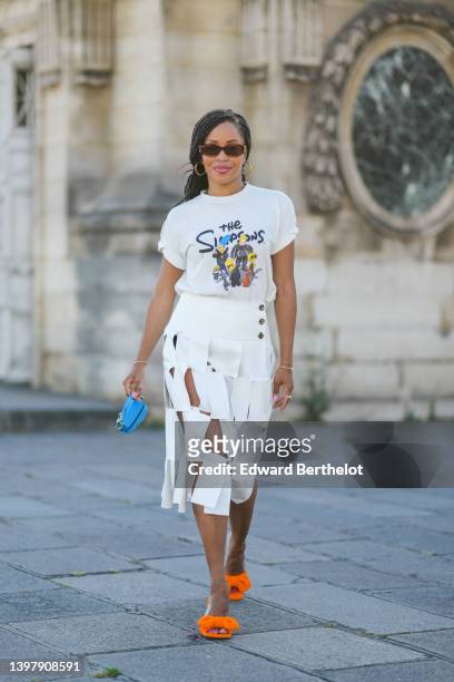Ellie Delphine wears brown sunglasses, gold earrings, a white with black and blue and yellow t-shirt with The Simpsons print pattern, gold bracelets,...
