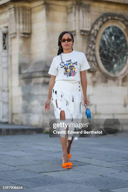 Ellie Delphine wears brown sunglasses, gold earrings, a white with black and blue and yellow t-shirt with The Simpsons print pattern, gold bracelets,...