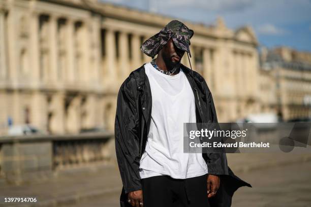 Cheick Bamba wears a gray / khaki / black military print pattern cap, a black nylon oversized jacket, a white oversized t-shirt, black flared pants,...