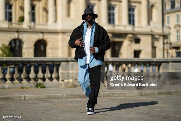 Cheick Bamba wears a silver large chain necklace , a black nylon bob hat, a white oversized t-shirt, a black denim jacket, a blue faded denim shirt,...