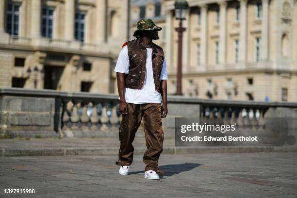 Cheick Bamba wears a beige / khaki / brown military print pattern nylon bob hat, a silver large chain necklace, a white oversized t-shirt, a dark...