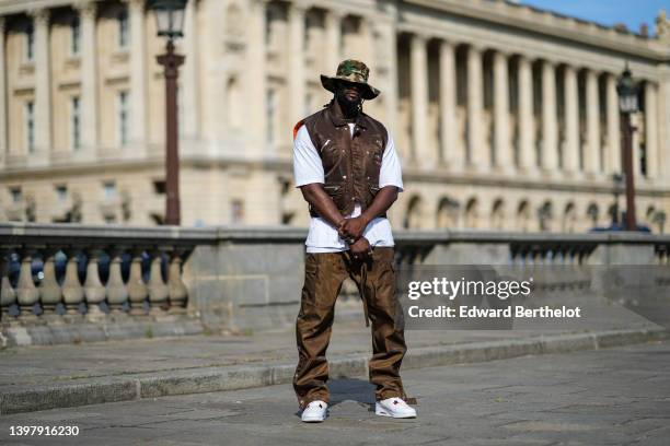 Cheick Bamba wears a beige / khaki / brown military print pattern nylon bob hat, a silver large chain necklace, a white oversized t-shirt, a dark...