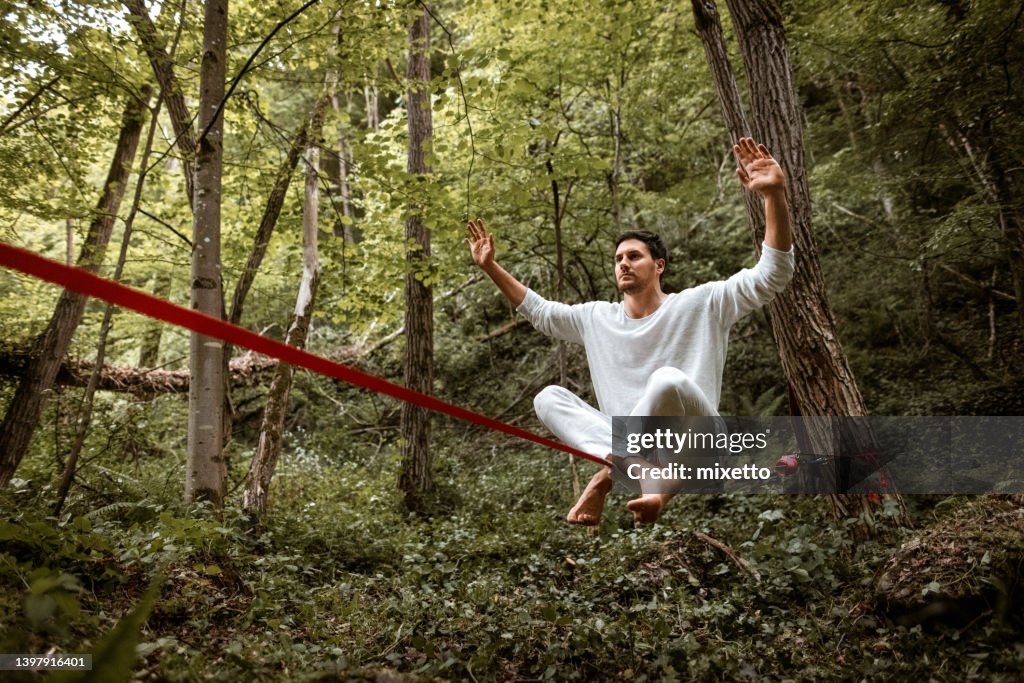 Junger Mann in einem weißen Pyjama-ähnlichen Anzug, der auf einer Slackline in einem Wald sitzt