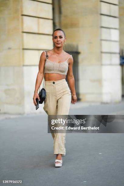 Alice Belaïdi wears gold oversized earrings, gold chain pendant necklaces, a beige cut-out pattern / V-neck / cropped tank-top, a black shiny leather...