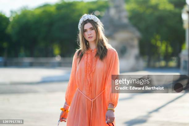 Zita d'Hauteville wears a white latte with blue embroidered flower pattern / braided silk headband, gold earrings, an orange ruffled / ribbed / long...