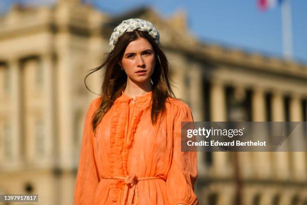 Zita d'Hauteville wears a white latte with blue embroidered flower pattern / braided silk headband, gold earrings, an orange ruffled / ribbed / long...