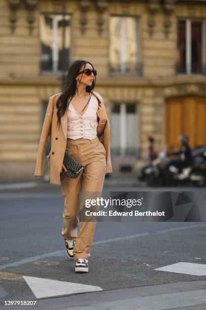 Gabriella Berdugo wears brown sunglasses, a white denim V-neck / buttoned / sleeveless gilet, a beige denim with pale pink embroidered flower...