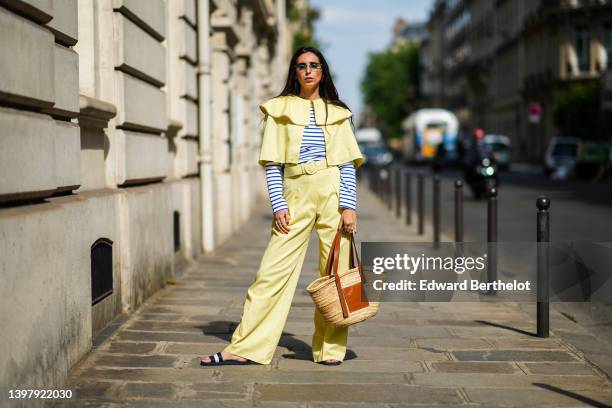 Gabriella Berdugo wears blue sunglasses, a navy blue and white striped print pattern long sleeves t-shirt, a yellow cropped cloak, matching yellow...